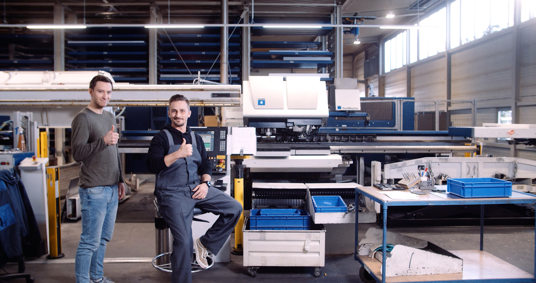 Ein Mitarbeiter eines Metallverarbeitungsbetriebs und der Protagonist des Laserhub-Werbefilms stehen stolz vor einer großen Maschine und zeigen den Daumen nach oben