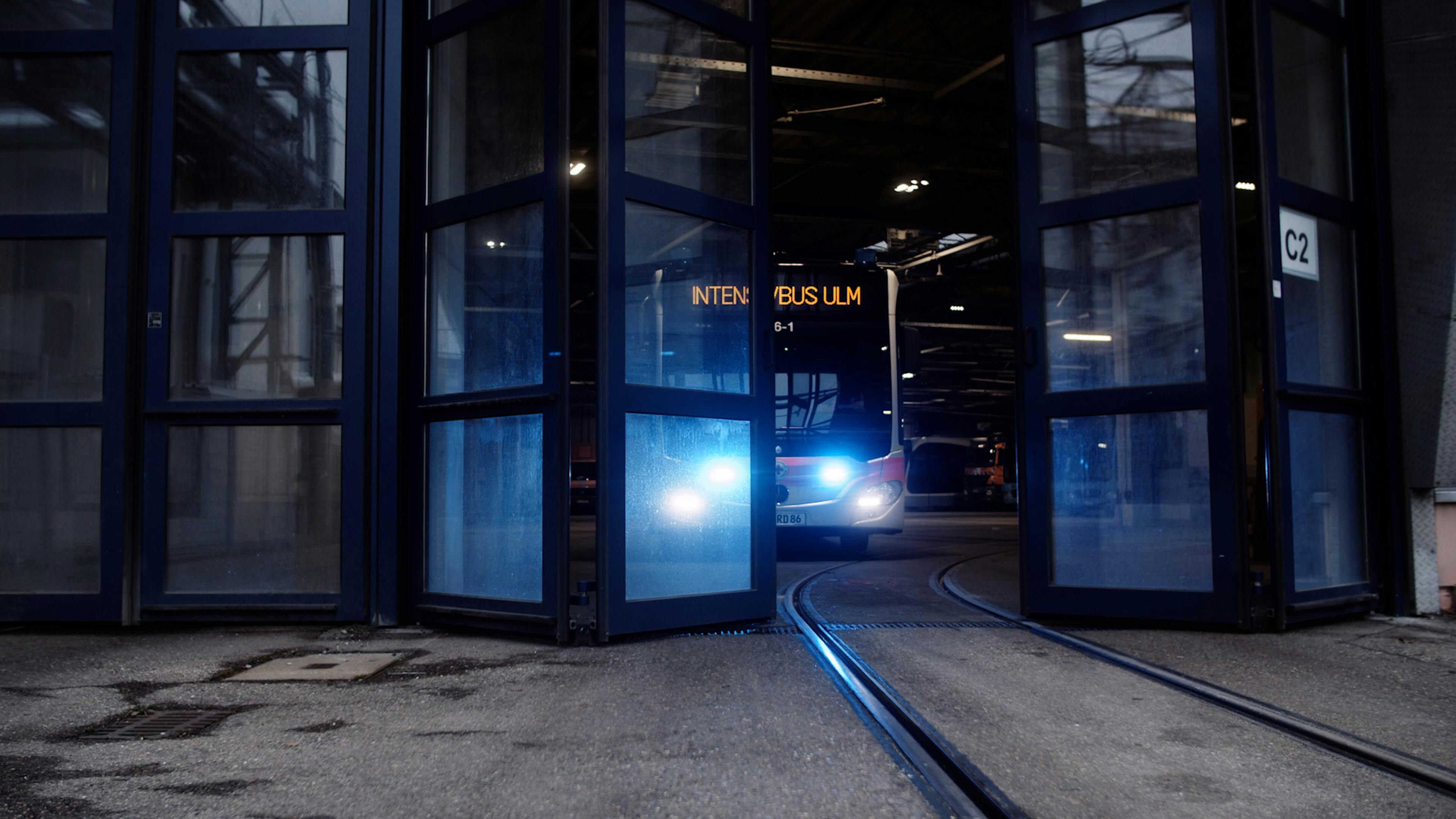 Ein teilweise durchsichtiges Tor öffnet sich, ein Linienbus mit Blaulicht und er Aufschrift "Intensivbus Ulm" fährt heraus