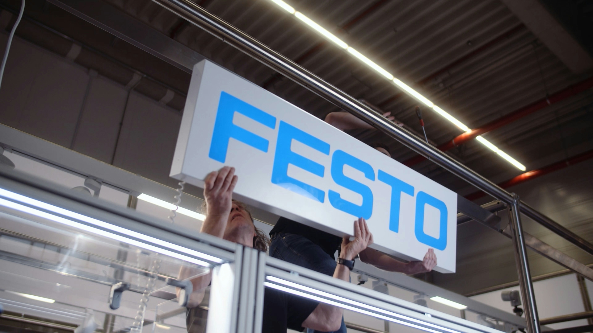 Zwei Mechaniker bringen eine Tafel mit dem Aufdruck des Festo Logos am Messestand an